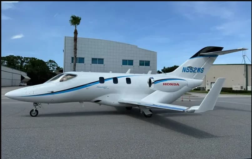 A side view of the white HondaJet HA-420, registration N552WS, with blue accents, parked on an airfield next to a hangar.