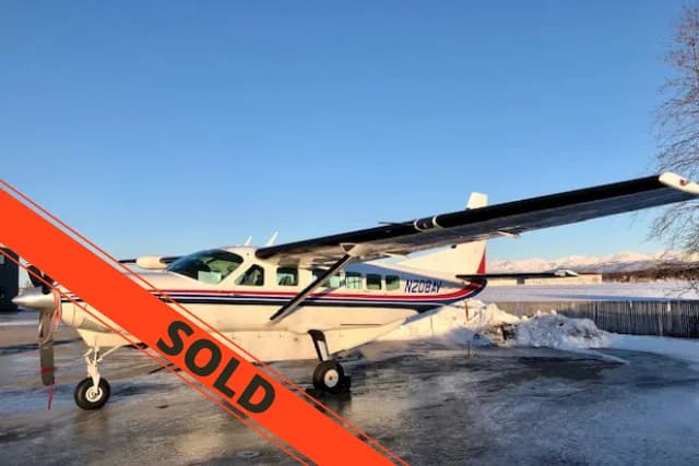 A white Cessna 208 Caravan with blue and red stripes is parked on an icy ramp with snow and mountains in the background under a clear blue sky.