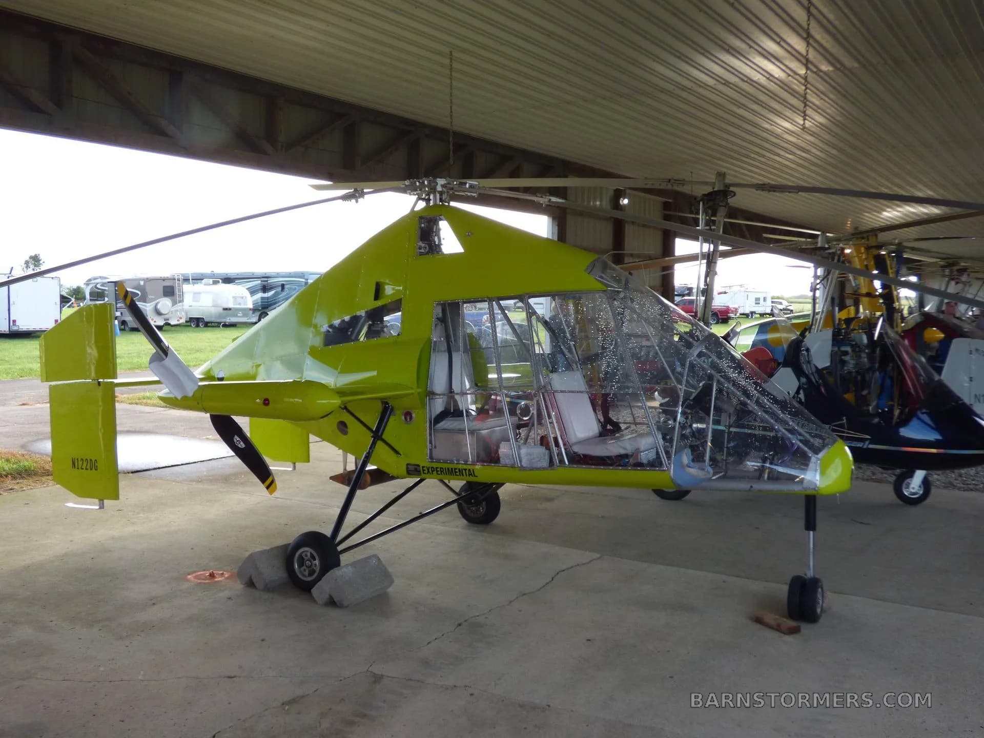 A bright green gyrocopter with its canopy open, parked inside a hangar with other aircraft and RVs visible outside.