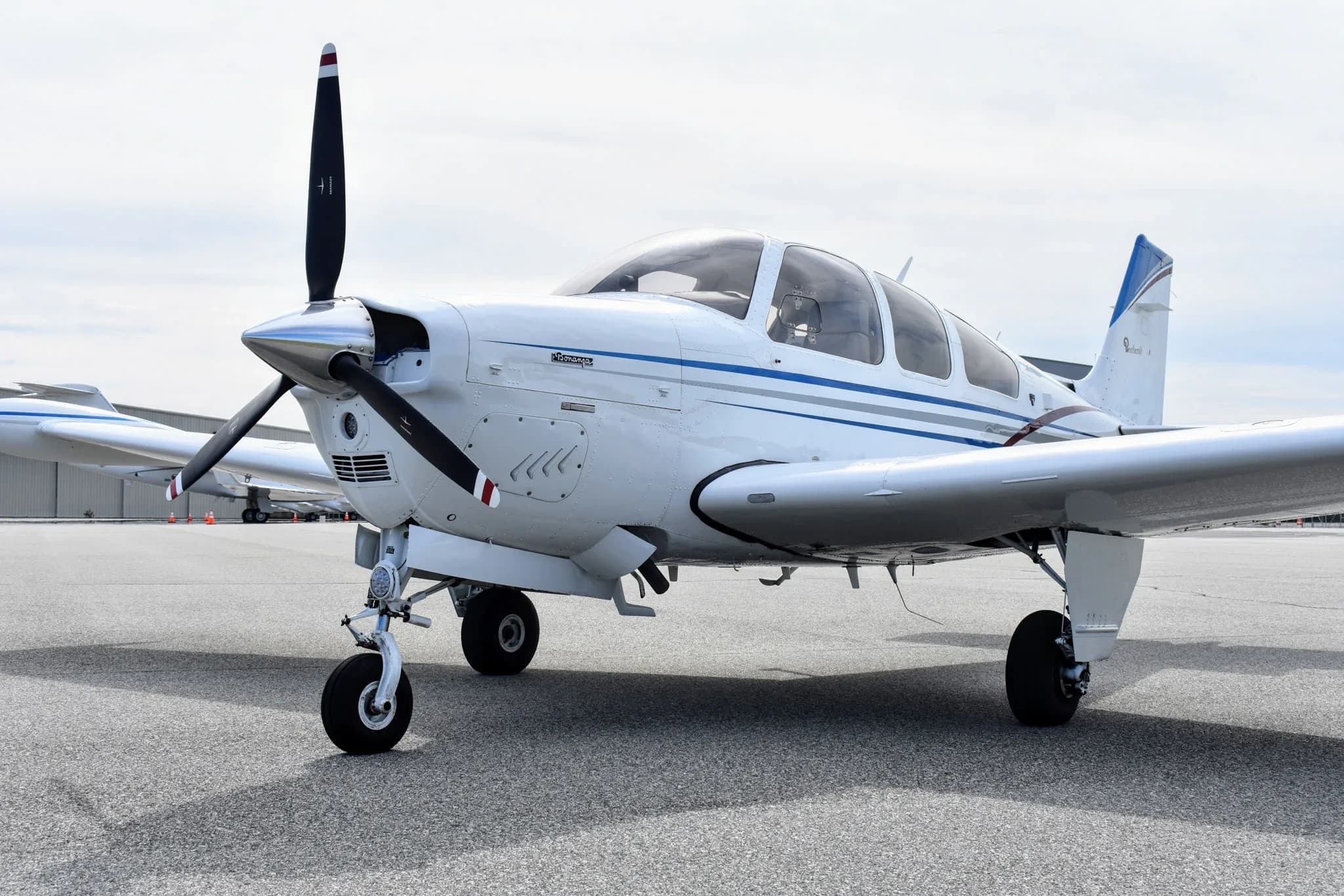 An angled front view of a white Beechcraft Bonanza F33A with blue and maroon stripes, parked on an asphalt tarmac.