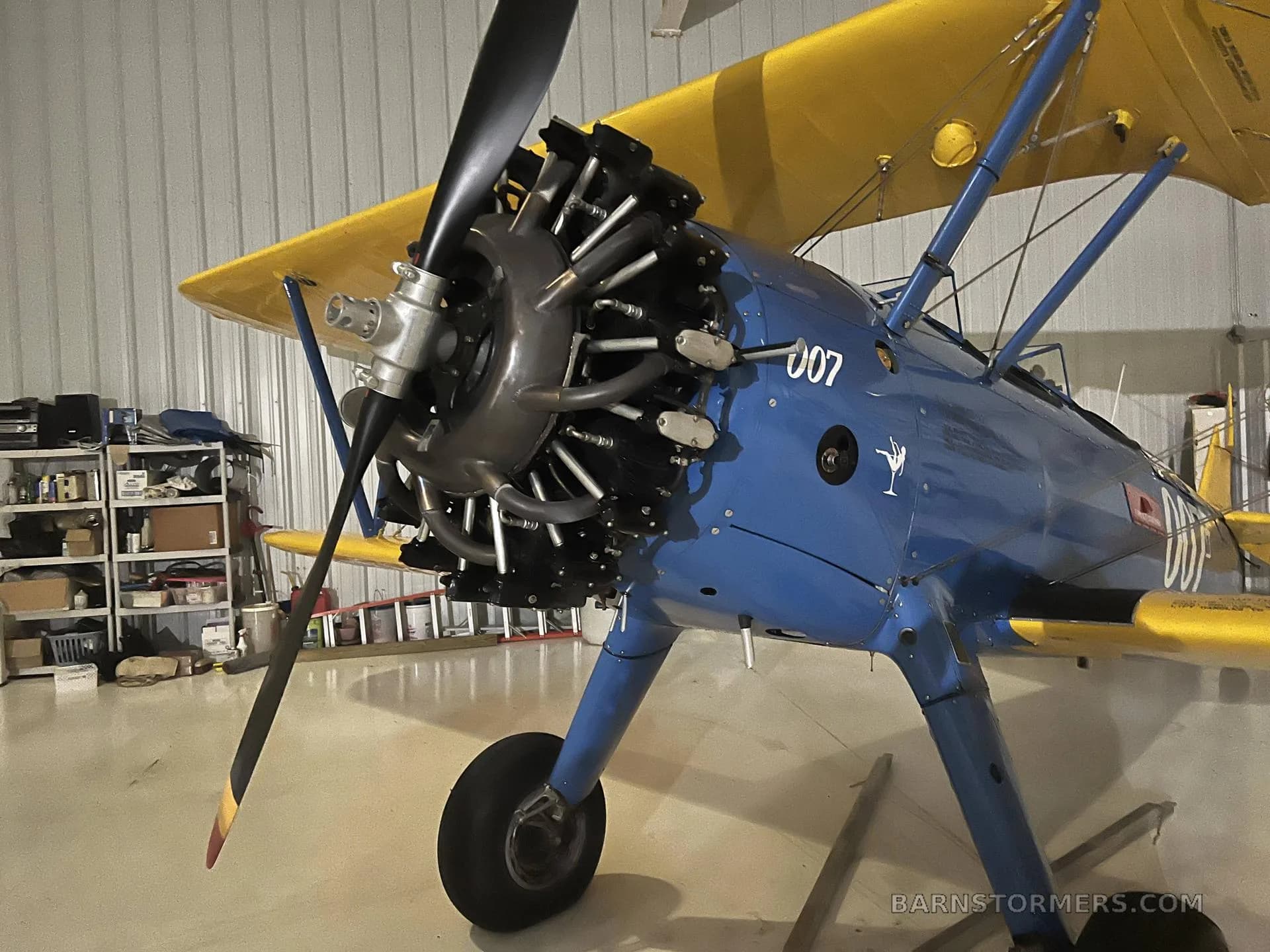 A close-up view of the radial engine and propeller of a blue and yellow biplane inside a hangar.