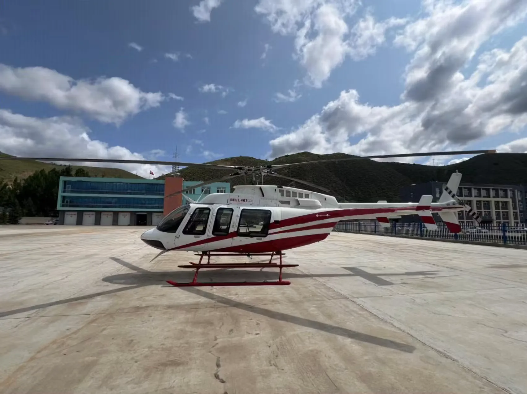 A white and red Bell 407 GXi helicopter parked on a concrete pad with buildings and mountains in the background.