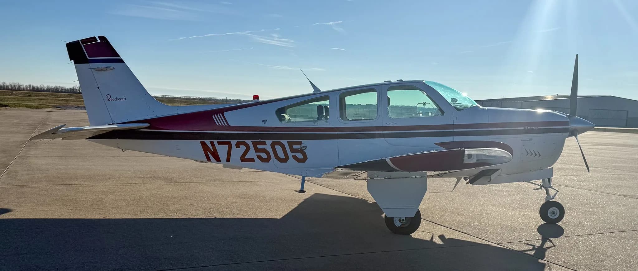 Side-left view of a white Beechcraft Bonanza with red and blue stripes, showing registration N72505, on an airfield with sun flare.