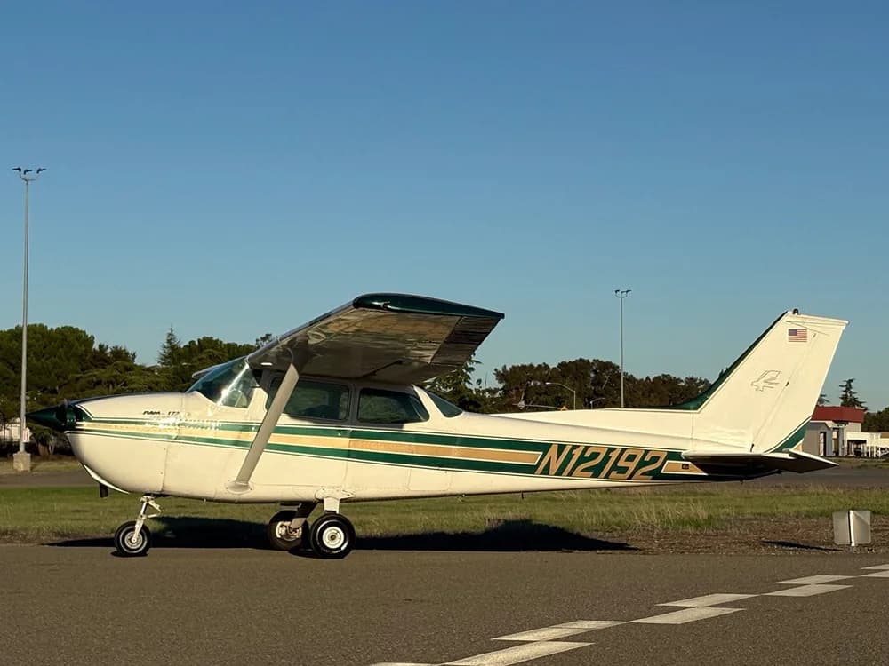A white Cessna 172 with green and gold stripes parked on an airfield next to a grassy area.