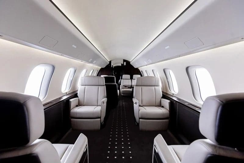 The luxurious cabin interior of a business jet featuring light grey leather seats arranged in a club configuration with a dark carpet.