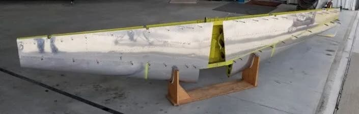 A partially assembled aircraft wing section, showing its metal skin and internal structure, on wooden stands.