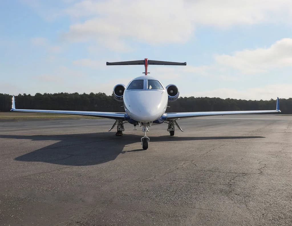 A white and blue Embraer Phenom 300 business jet is parked on an asphalt runway, viewed directly from the front.
