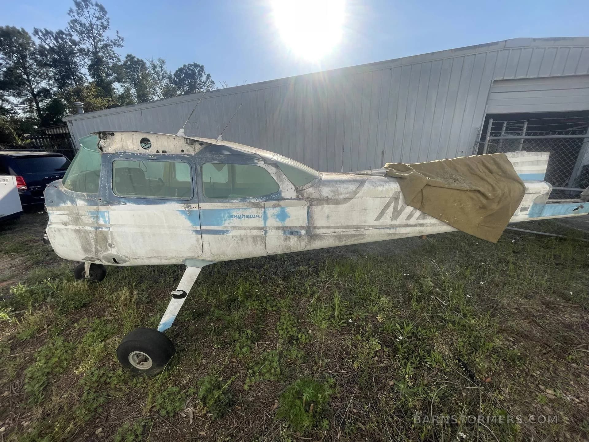 A side view of a weathered, partially disassembled Cessna Skyhawk aircraft with blue and white paint, parked on grass next to a building.