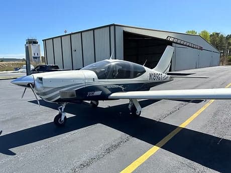 A Trinidad GT aircraft is parked on the tarmac in front of a hangar.