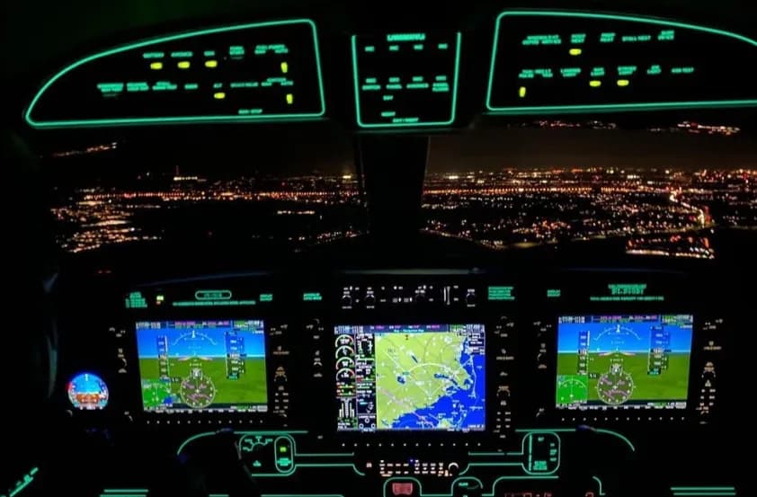 The illuminated cockpit of an aircraft at night, showing multiple display screens and city lights below.