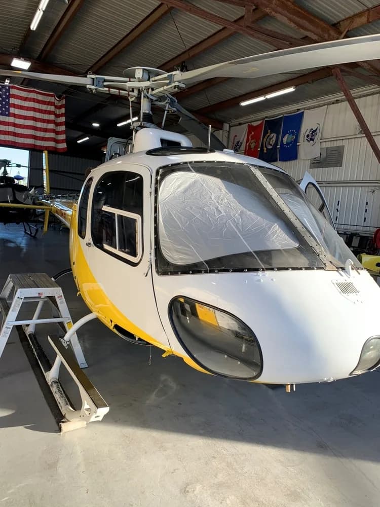 The front of a white and yellow Eurocopter AS350 B3 helicopter, N211FN, is seen inside a hangar with a sunshade in the windshield.