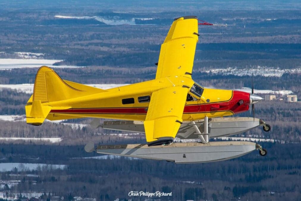 Yellow and red floatplane in flight over a forest and frozen lake.