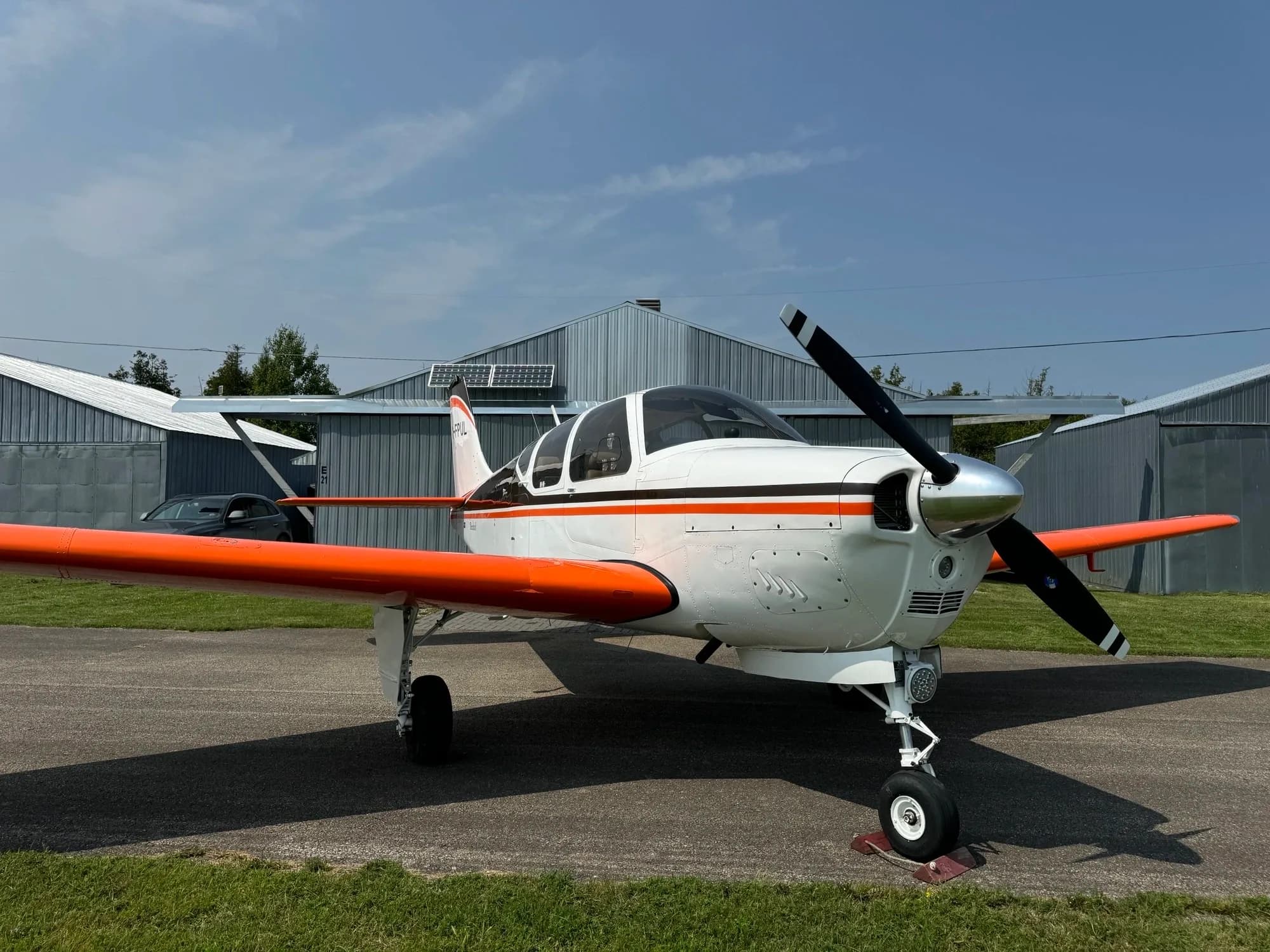 A white single-engine propeller aircraft with bright orange wings and tail is parked on an asphalt ramp in front of hangars under a clear sky.