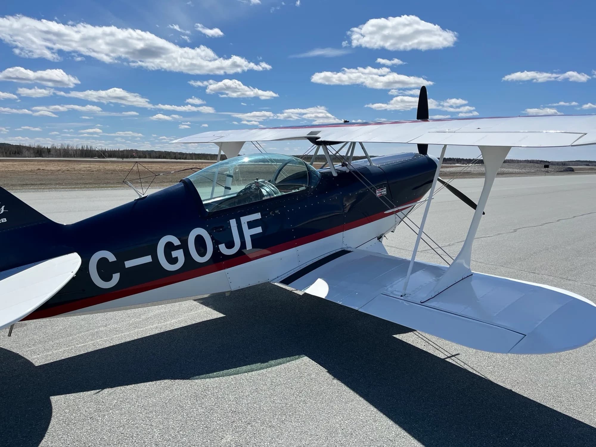 A side view of the dark blue and white Pitts biplane C-GOJF on an asphalt runway with a clear blue sky.