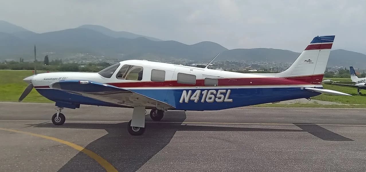 A Piper Saratoga II TC aircraft with white, blue, and red paint scheme on a tarmac with mountains in the background.