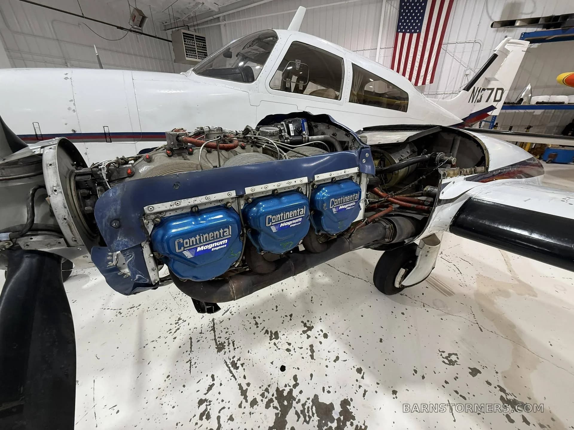 A side view of a white single-engine aircraft with its Continental engine exposed for maintenance, showing blue valve covers, in a hangar with an American flag.