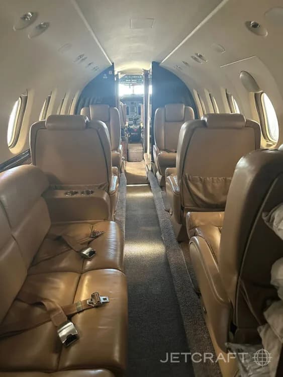 The interior cabin of a private jet with tan leather seats and an aisle leading to the cockpit.
