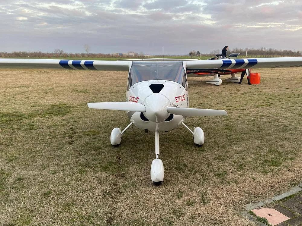 A white Pipistrel Sinus aircraft with blue wing stripes, viewed from the front on a grassy airfield under a cloudy sky.