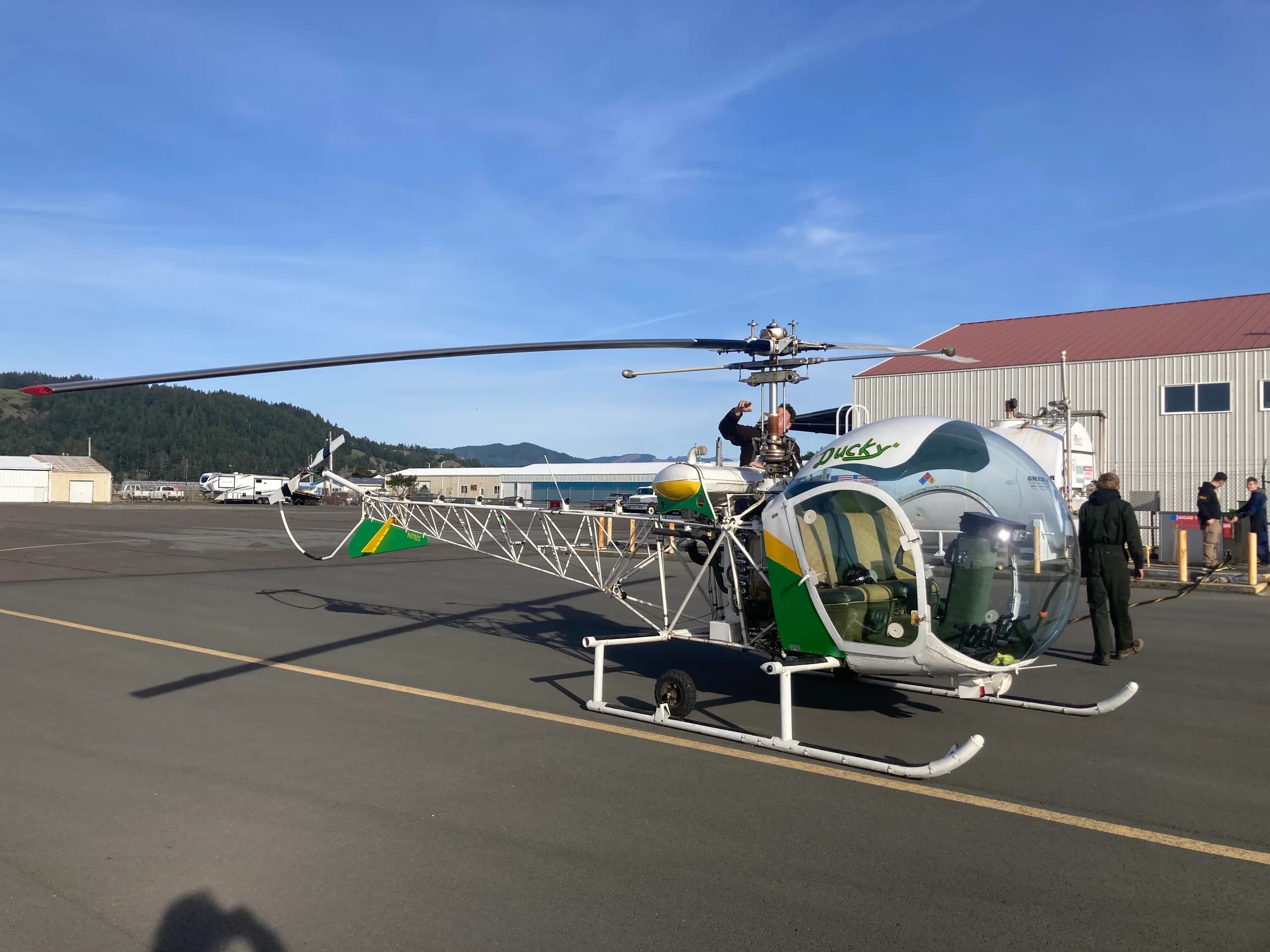A white, green, and yellow open-frame helicopter named 'Ducky' sits on an asphalt tarmac with people, buildings, and hills in the background under a clear sky.