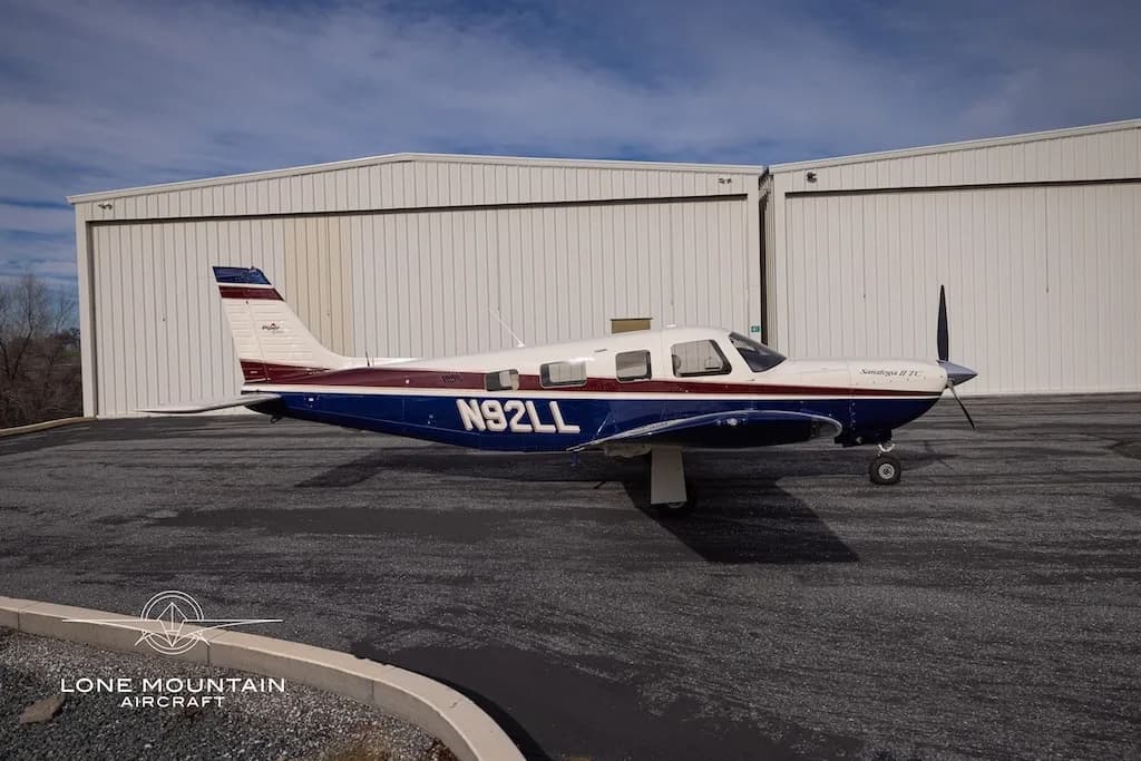 A side view of the Piper Saratoga II TC aircraft (N92LL) with blue and white paint scheme parked on an asphalt apron in front of hangars.