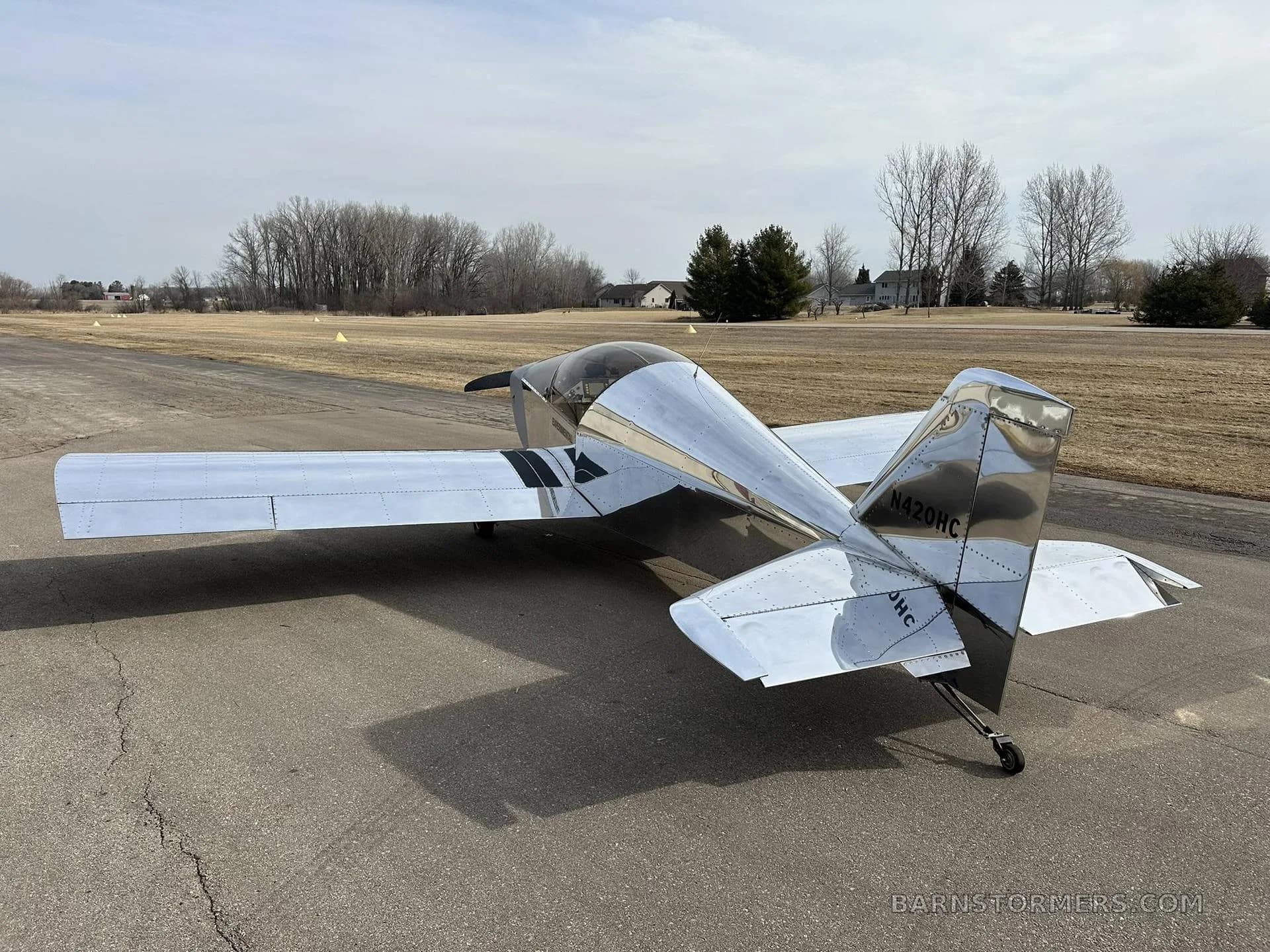 Rear-side view of a shiny silver experimental aircraft with "N420HC" on the tail, parked on an asphalt surface.
