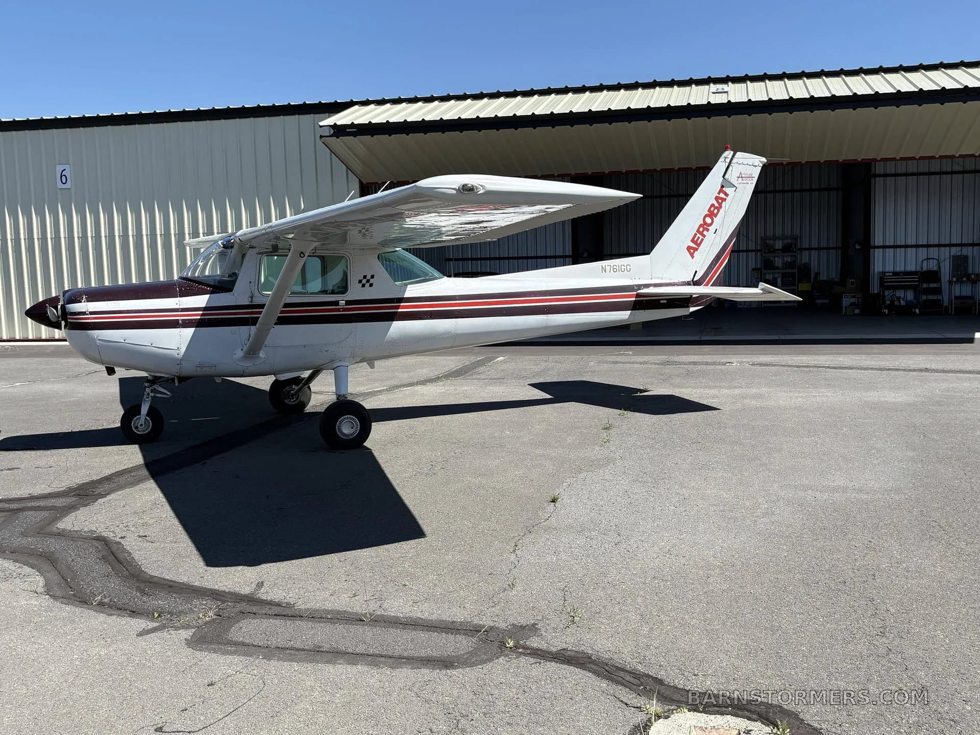 A white and maroon Cessna 150 Aerobat (N761GG) parked on an asphalt apron next to a hangar.
