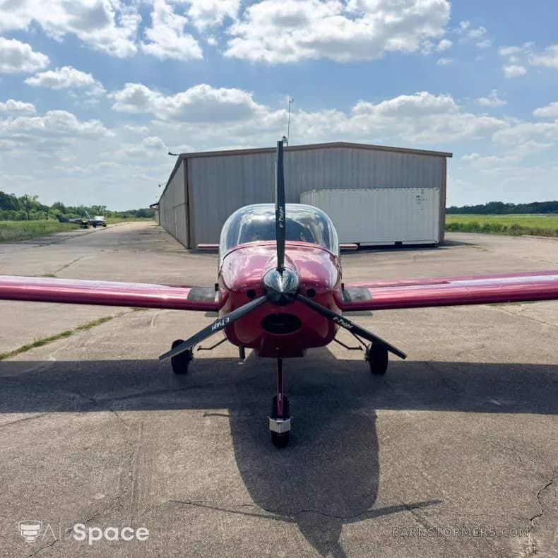 A head-on view of the red Vans RV-6 aircraft with a two-blade propeller, parked on a concrete tarmac in front of a hangar.