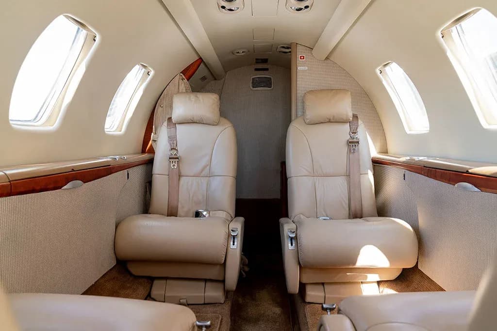 The interior cabin of a private jet, showing two rows of beige leather seats facing each other.