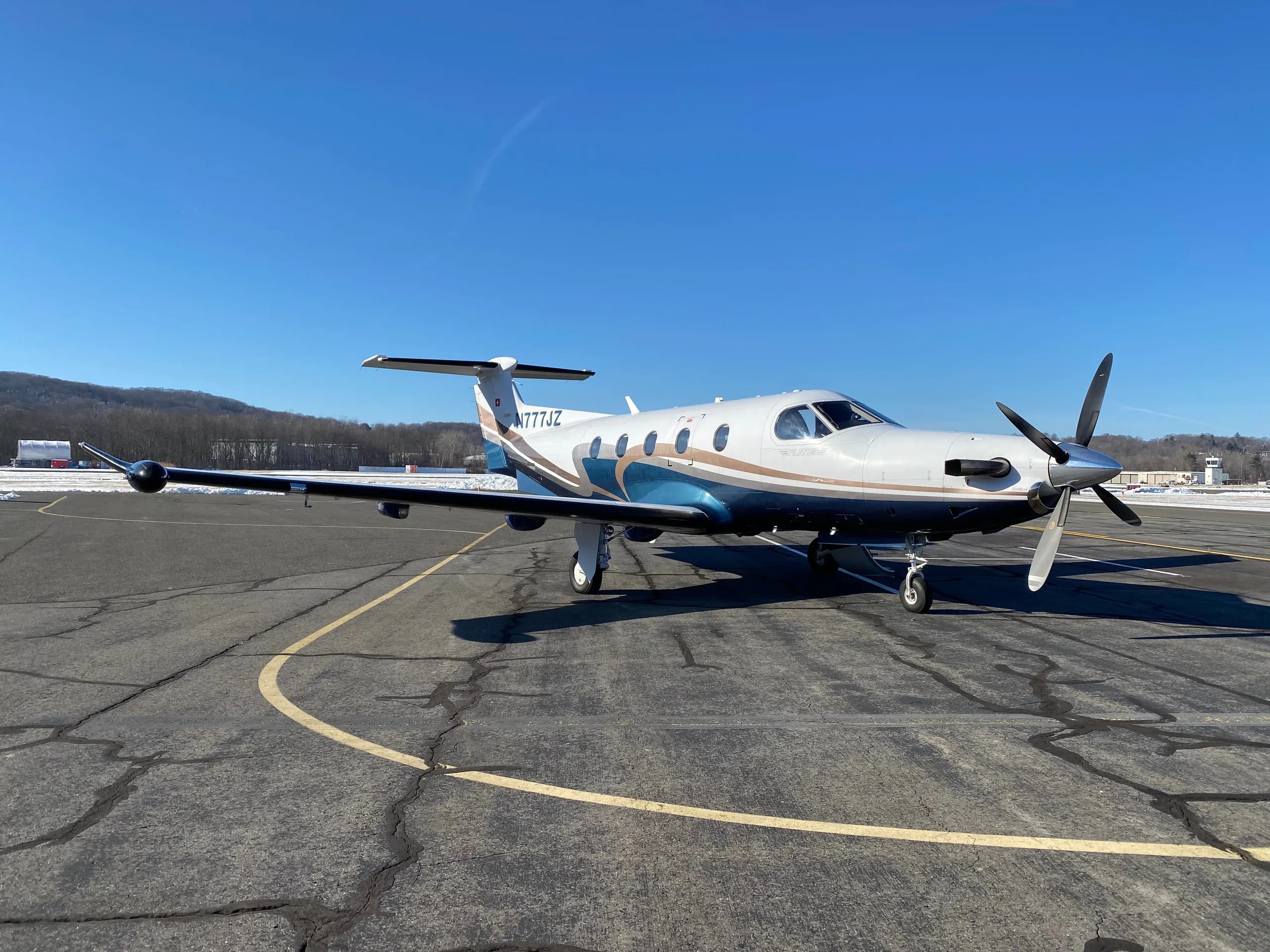 A Pilatus PC-12 aircraft with registration N777JZ is parked on the tarmac with a blue and brown paint scheme.