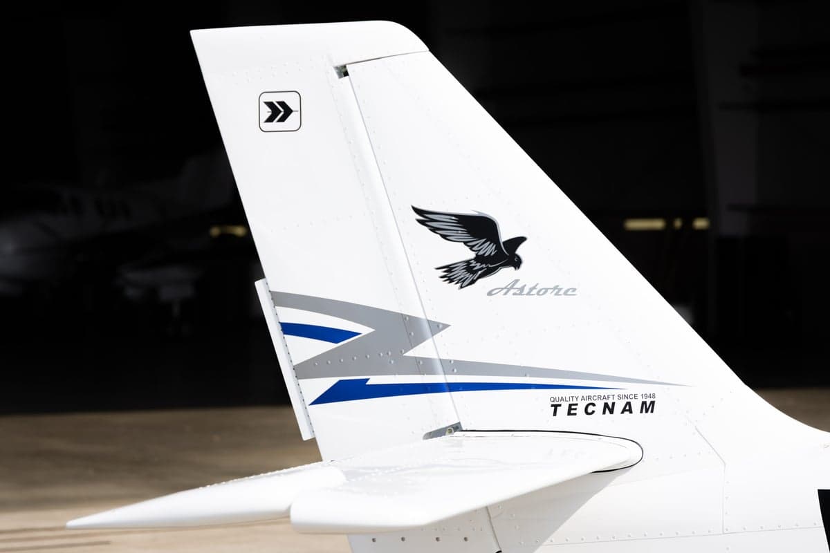 A close-up of the white tail of a Tecnam Astore aircraft, featuring a black bird logo and "Tecnam" text.