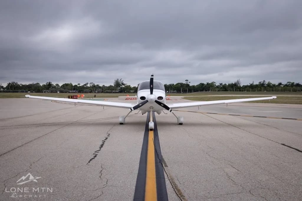A head-on view of a white Cirrus SR20 aircraft with orange and black stripes, registration N5886M, on an airfield runway centerline.