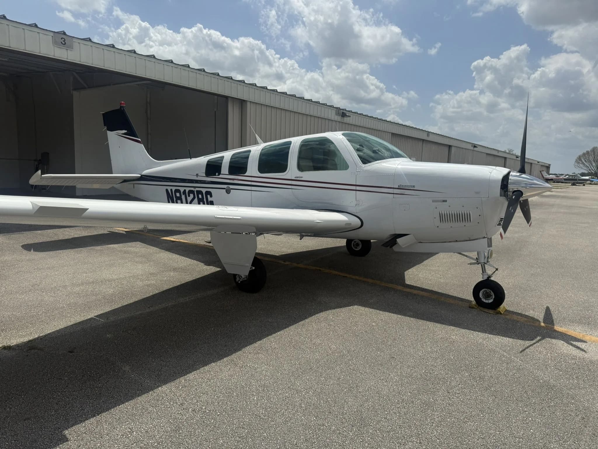 Front three-quarter view of a white Beechcraft Bonanza N812BG with dark red and black stripes on an airfield.