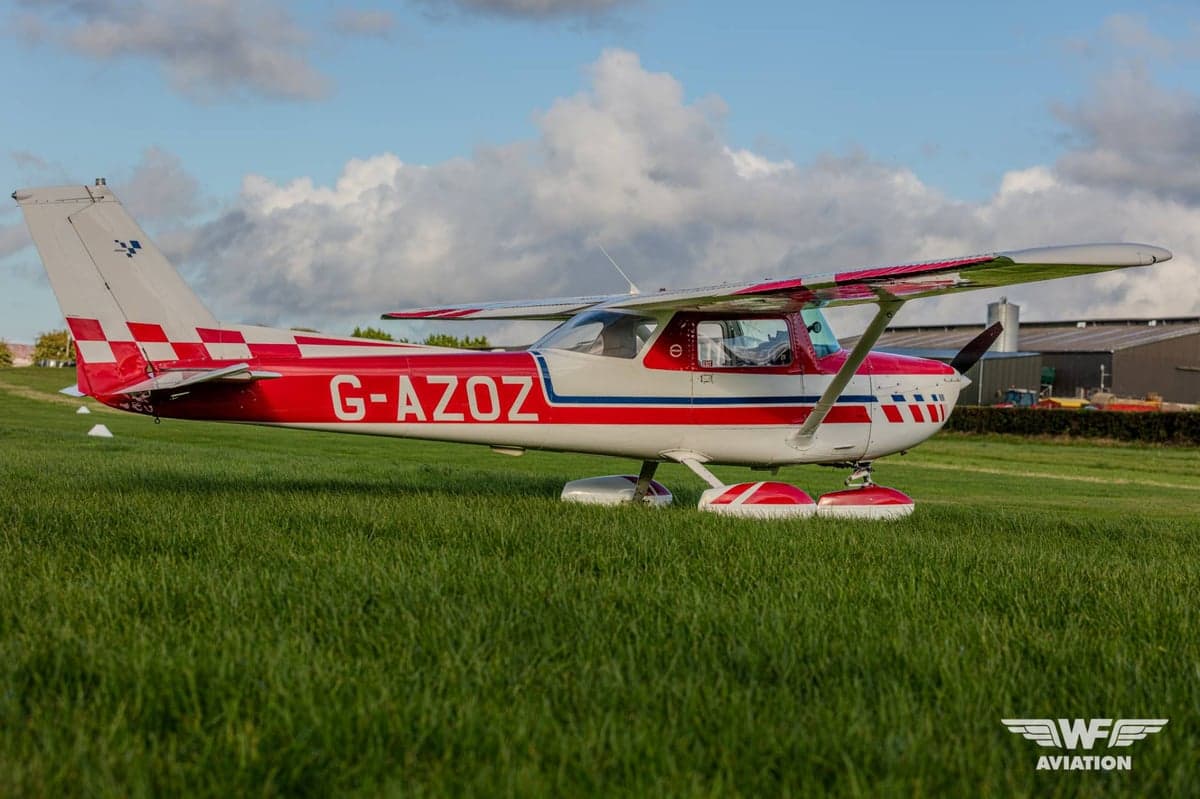 A red and white Cessna 150 Aerobat (G-AZOZ) with a checkered tail, parked on a grassy airfield under a cloudy sky.