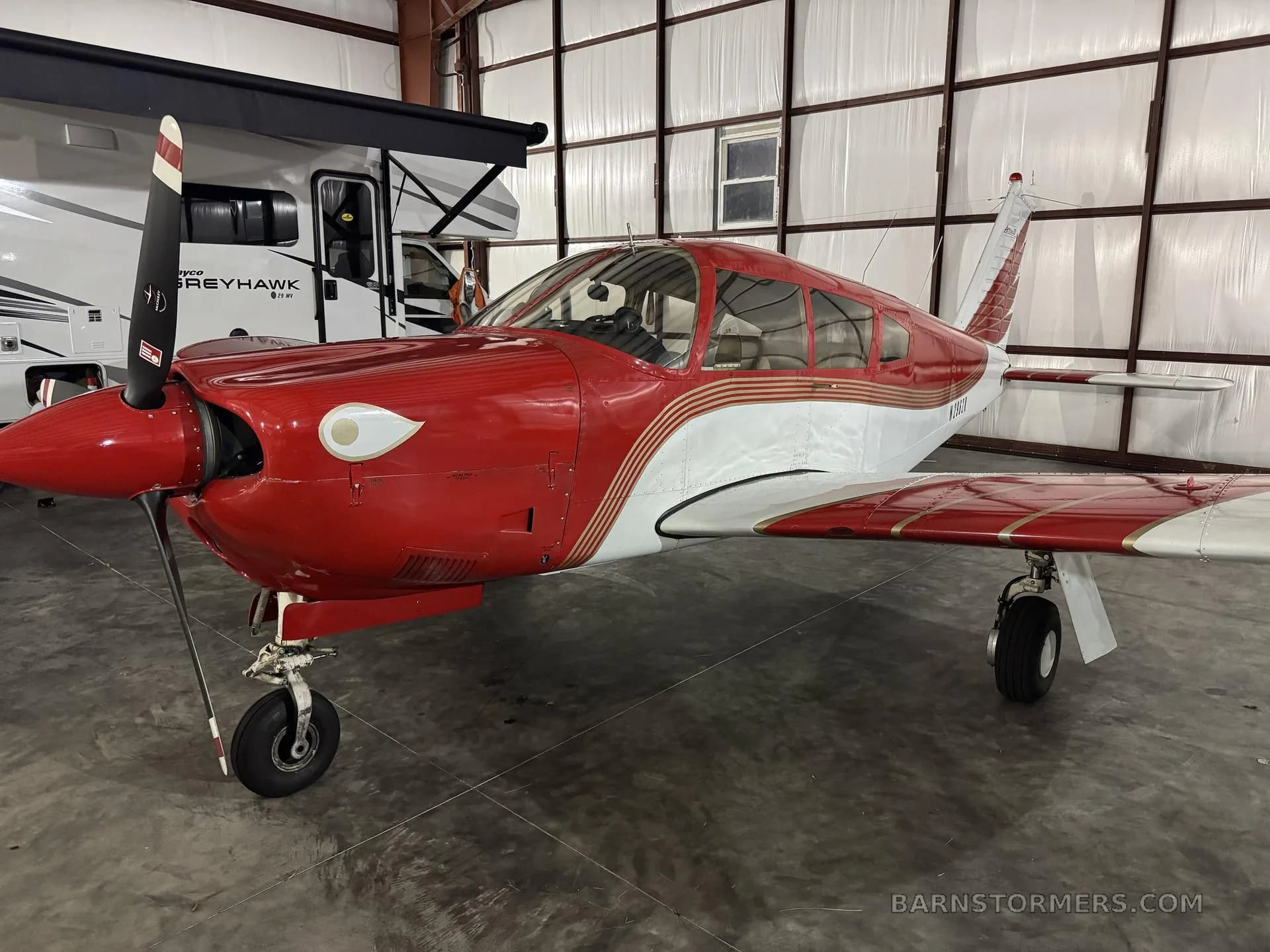 A red and white Mooney M20 series aircraft parked inside a hangar next to an RV.