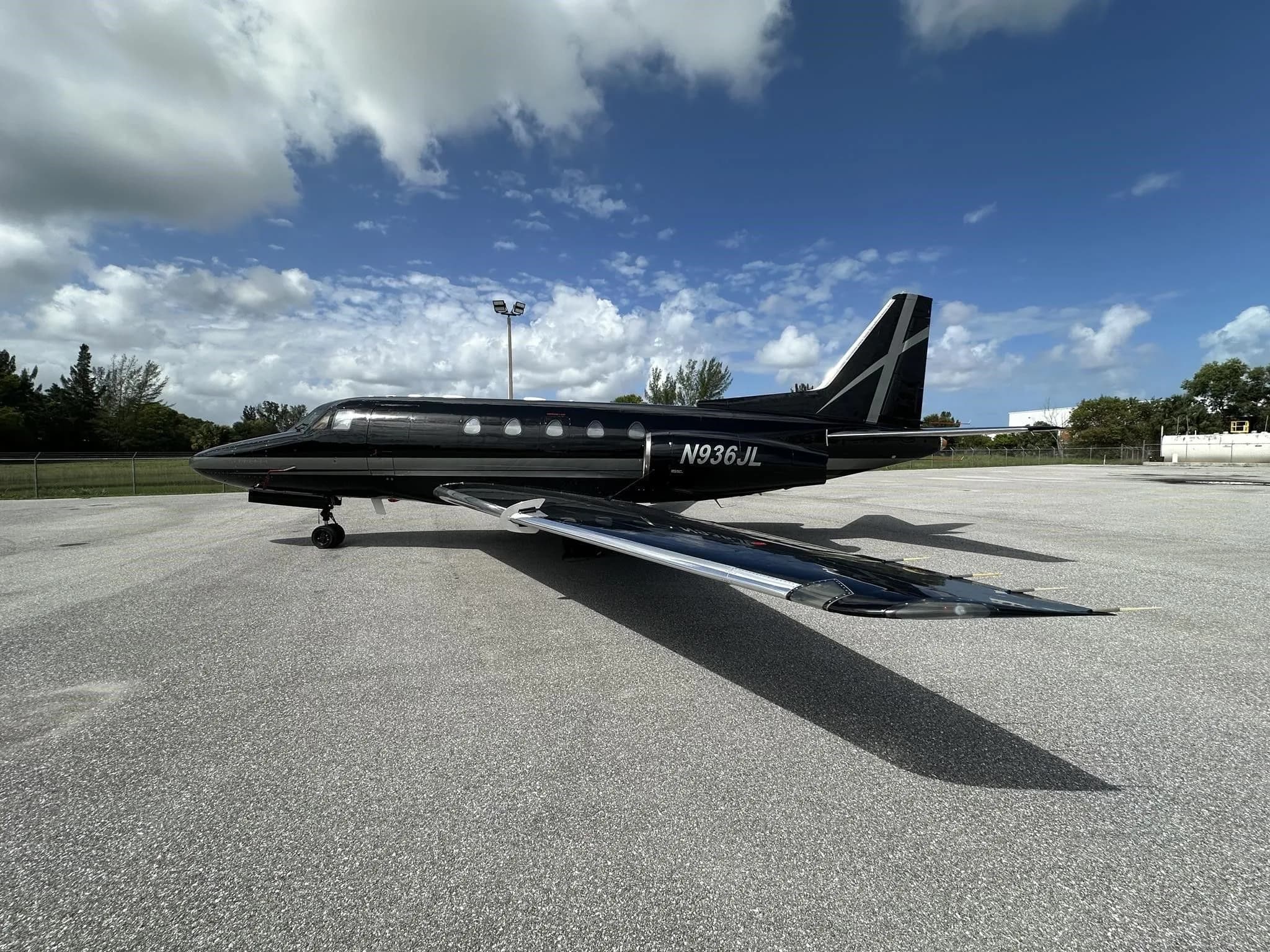 A black and silver business jet, N936JL, with its wings reflecting the sky, parked on an asphalt tarmac.