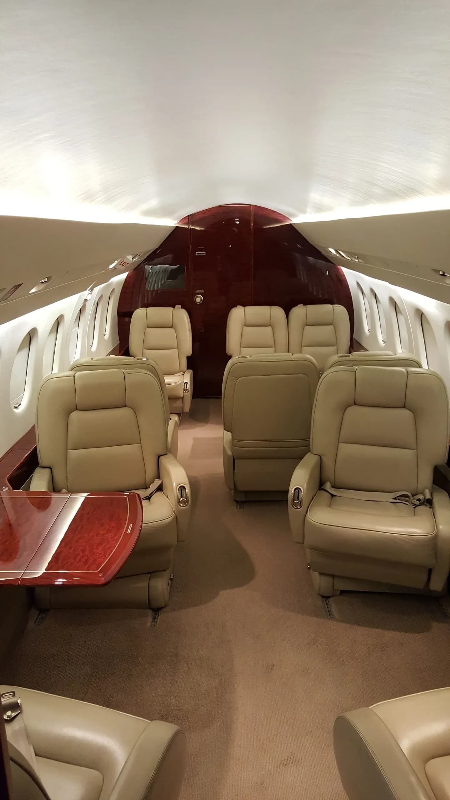 The spacious interior cabin of a private jet featuring beige leather seating and a polished wooden table.