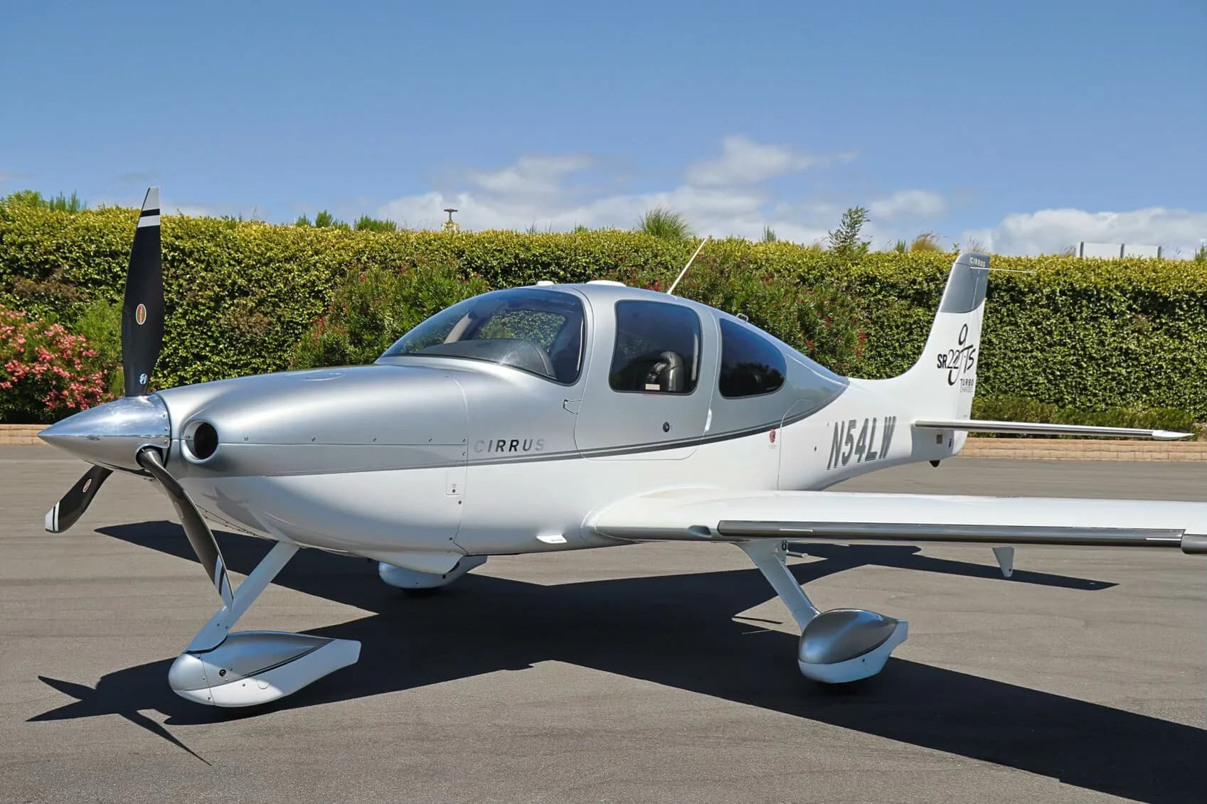 Front three-quarter view of a silver and white Cirrus SR22T (N54LW) on an tarmac, with green hedges in the background.