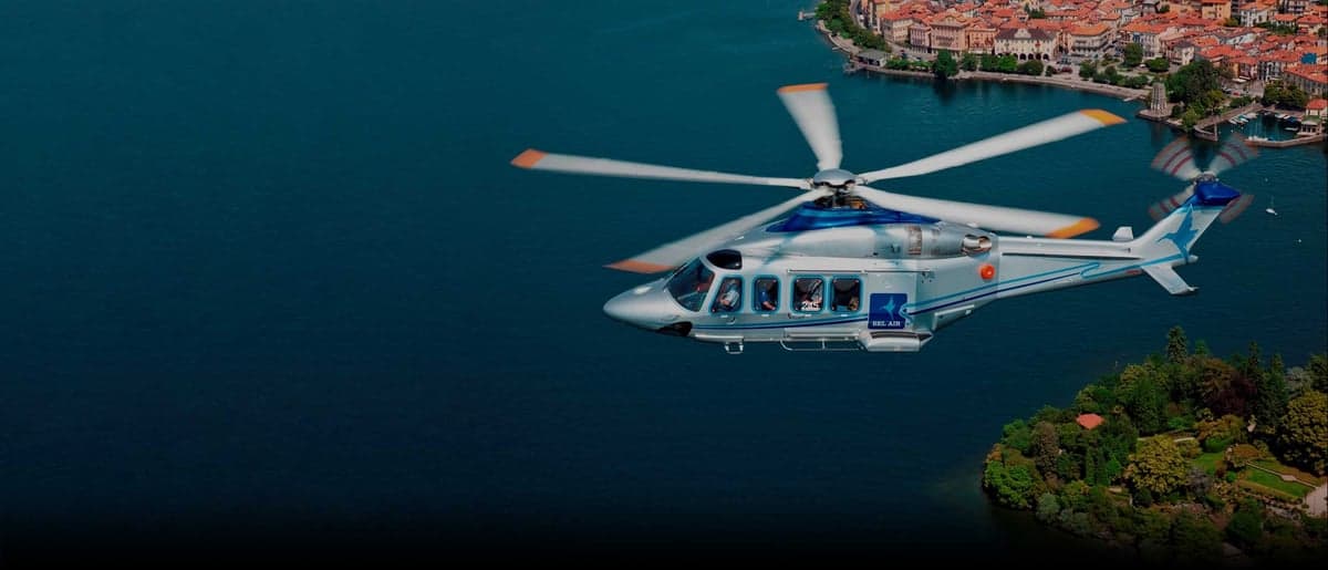 A silver and blue helicopter flies over a body of water near a town.