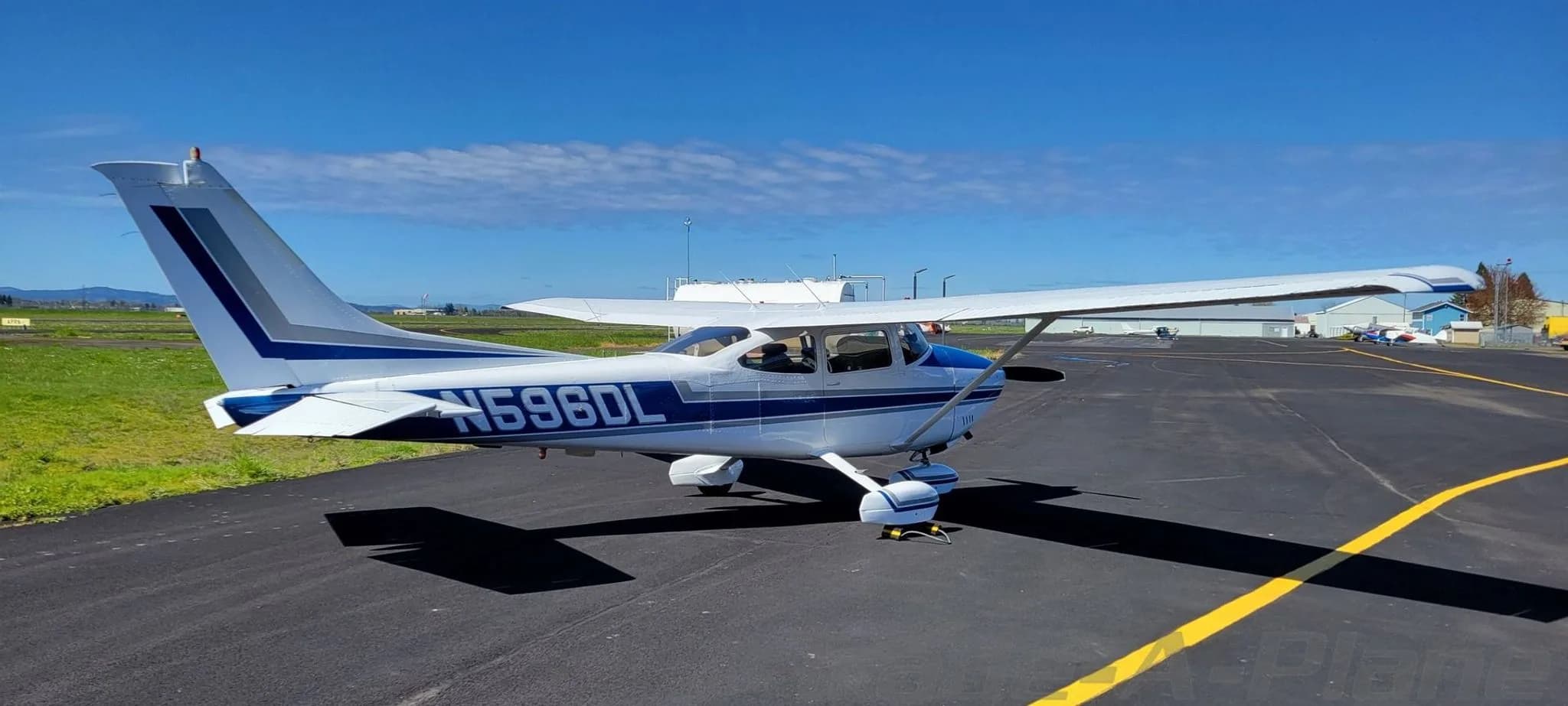 A white and blue Cessna 182 Skylane aircraft with registration N596DL parked on an airfield under a clear sky.