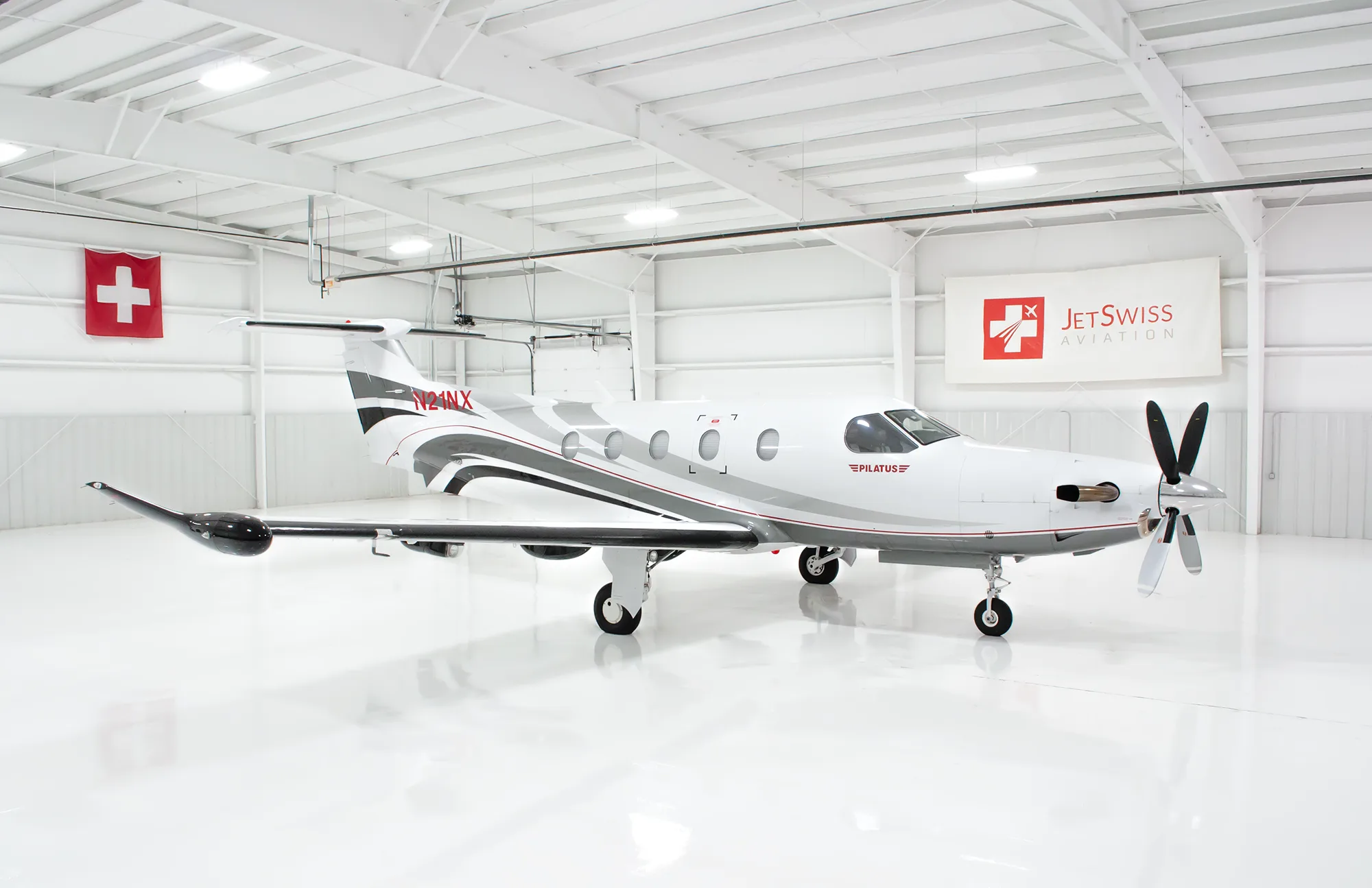 A white Pilatus PC-12 aircraft with gray and black stripes, registration N21NX, parked in a brightly lit hangar.