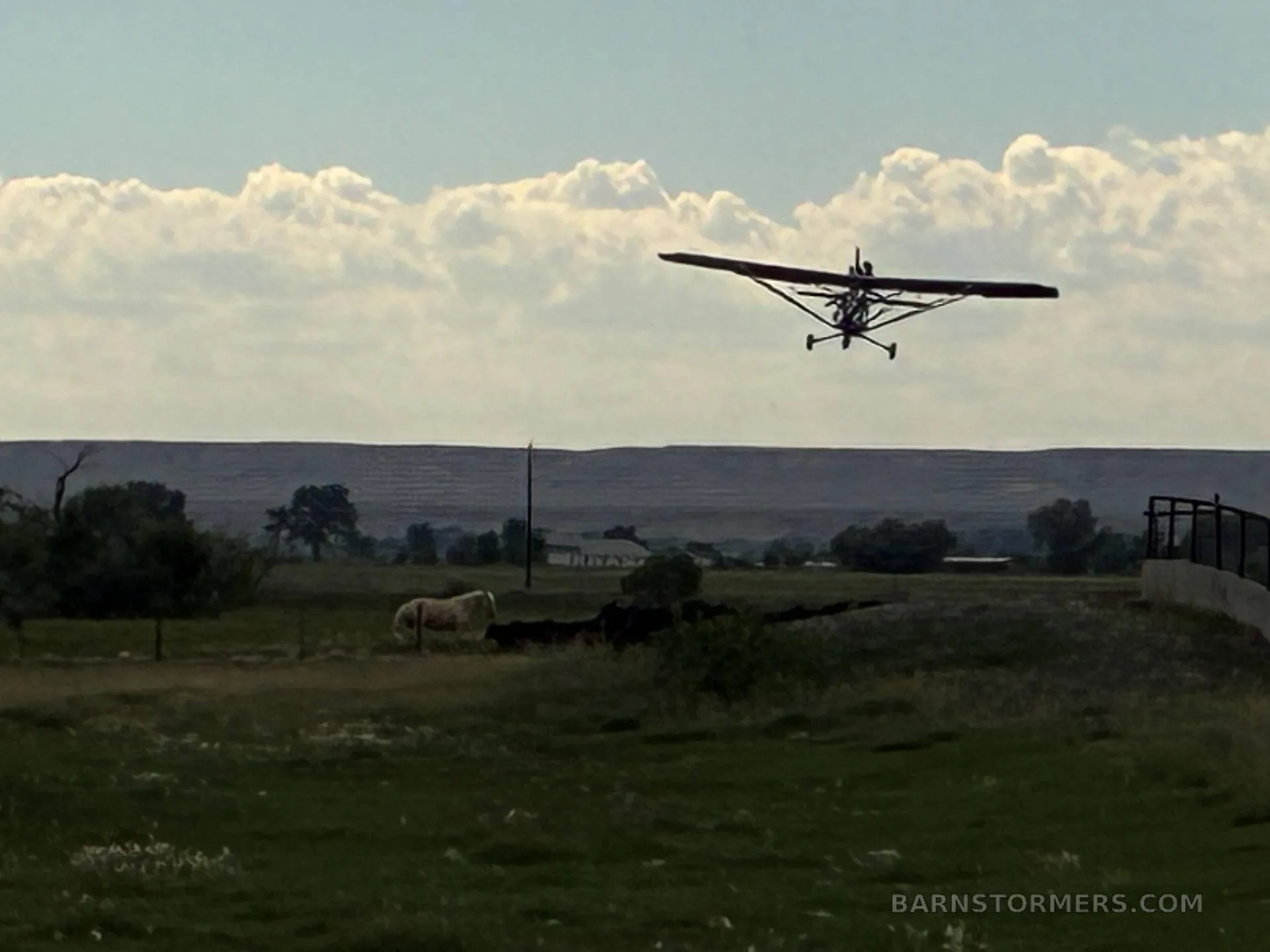 A red and blue ultralight aircraft in flight over a green field with distant hills and a cloudy sky.