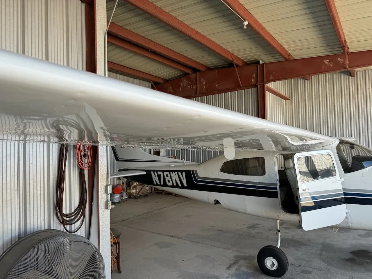 A white Cessna aircraft with blue and black stripes, registration N78WV, with its door open, parked in a hangar.