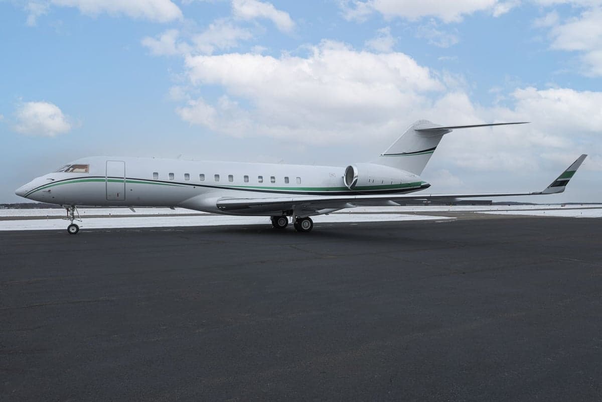 A white Bombardier Global business jet with green and black stripes parked on a snowy tarmac under a cloudy sky.