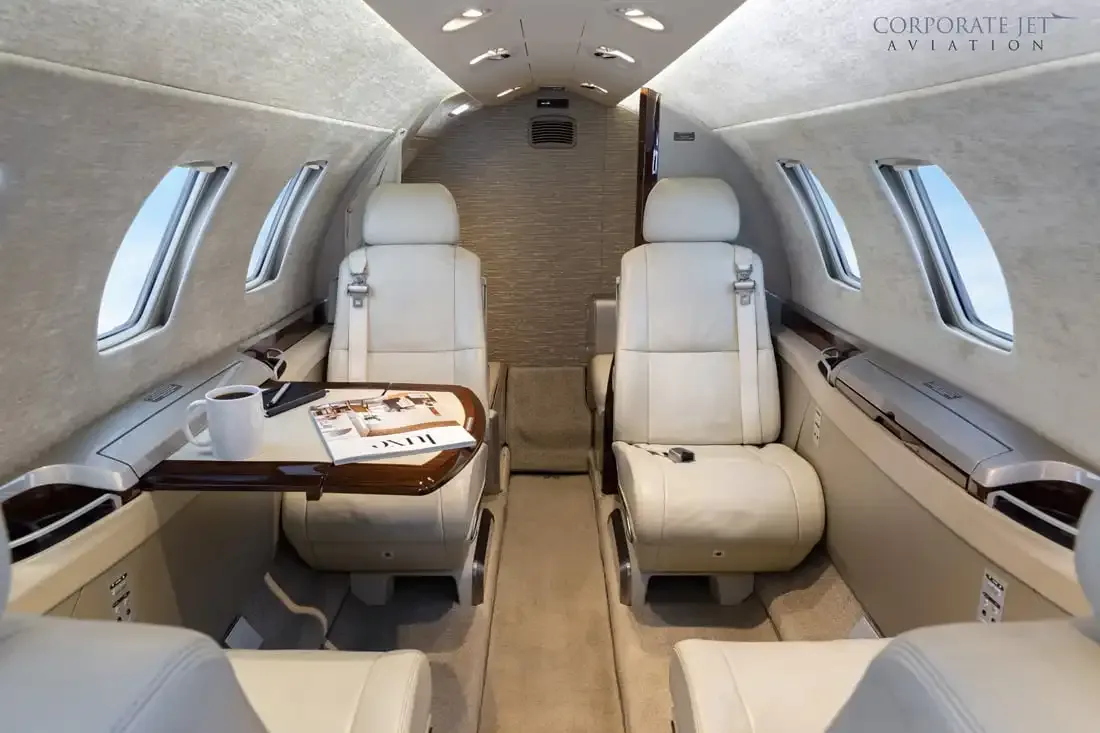 The interior of a private jet cabin with light-colored leather seats and a table with a magazine and coffee cup.