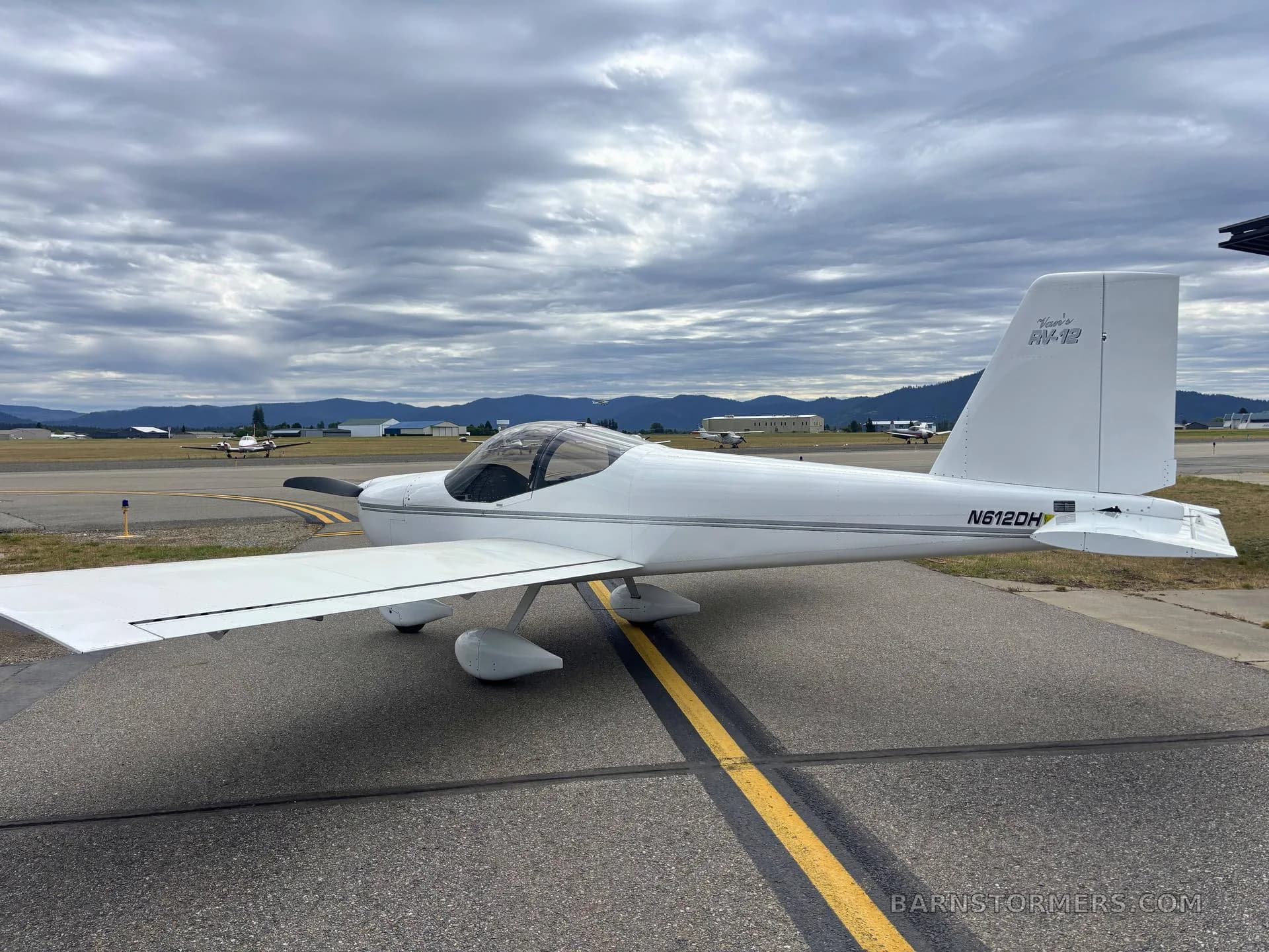 A white Vans RV-12 aircraft (N612DH) is parked on an airfield under a cloudy sky, viewed from the side.
