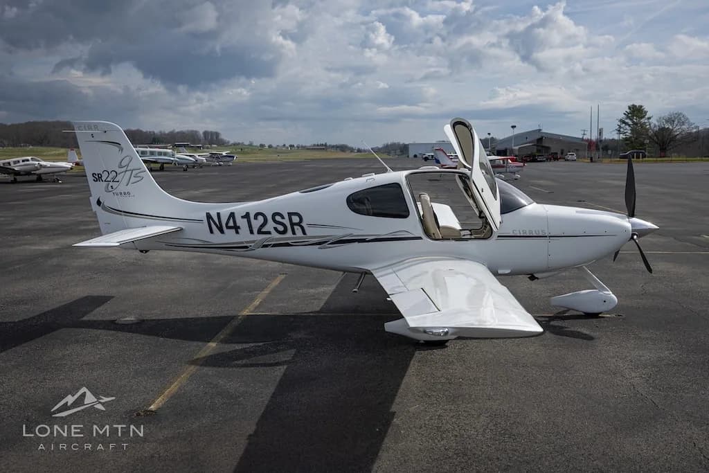A white Cirrus SR22T aircraft with open doors is parked on an asphalt runway with other planes in the distance.