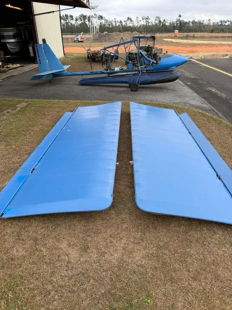 Two detached blue aircraft wings in the foreground, with the main blue ultralight aircraft body visible in the background near a hangar.