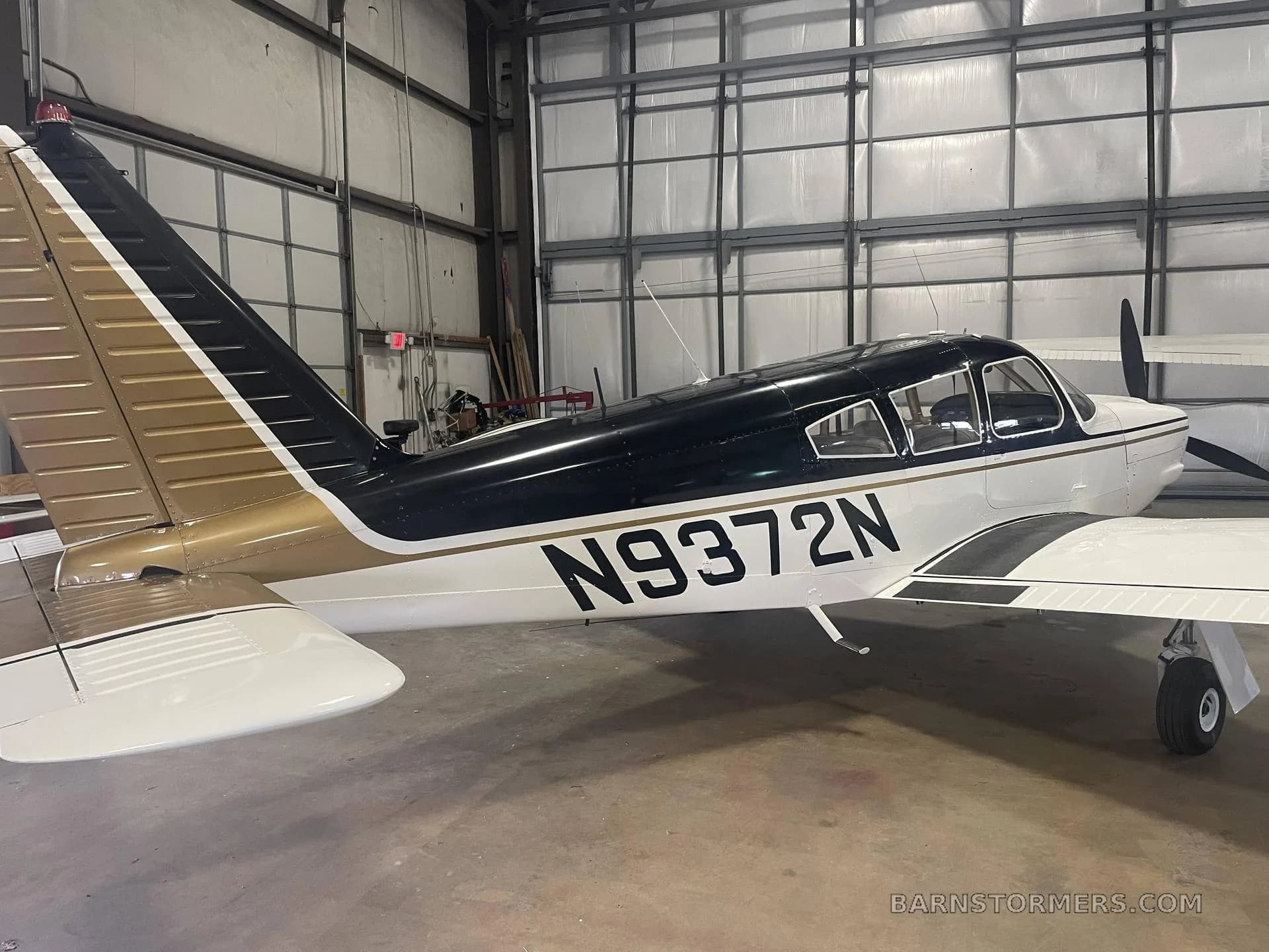 A white and black single-engine aircraft with a gold stripe, registration N9372N, parked in a hangar, viewed from the rear-left.