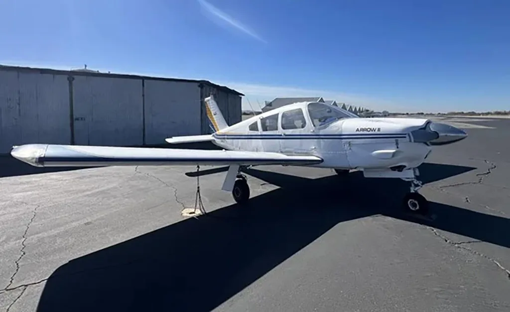 A white Piper Arrow II aircraft with blue and yellow stripes parked on an asphalt tarmac next to a hangar.