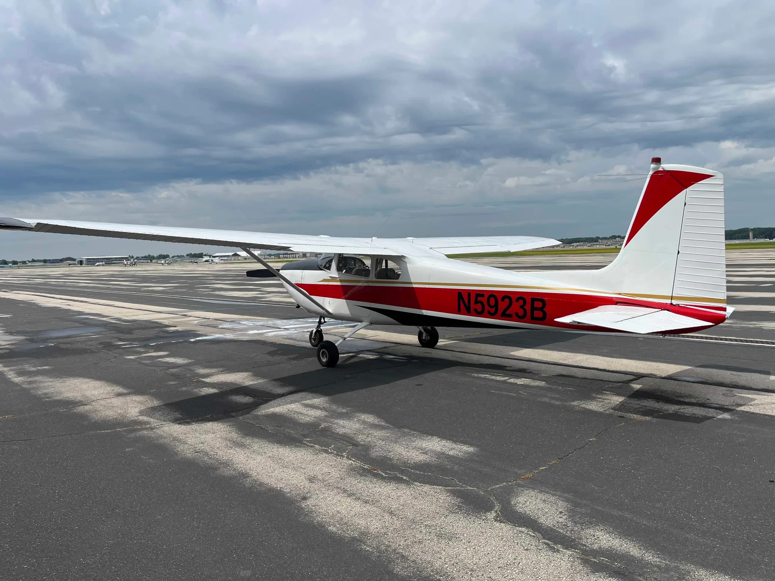 A side view of a white Cessna 182 aircraft with red and black stripes and registration N5923B, parked on an airport taxiway under a cloudy sky.